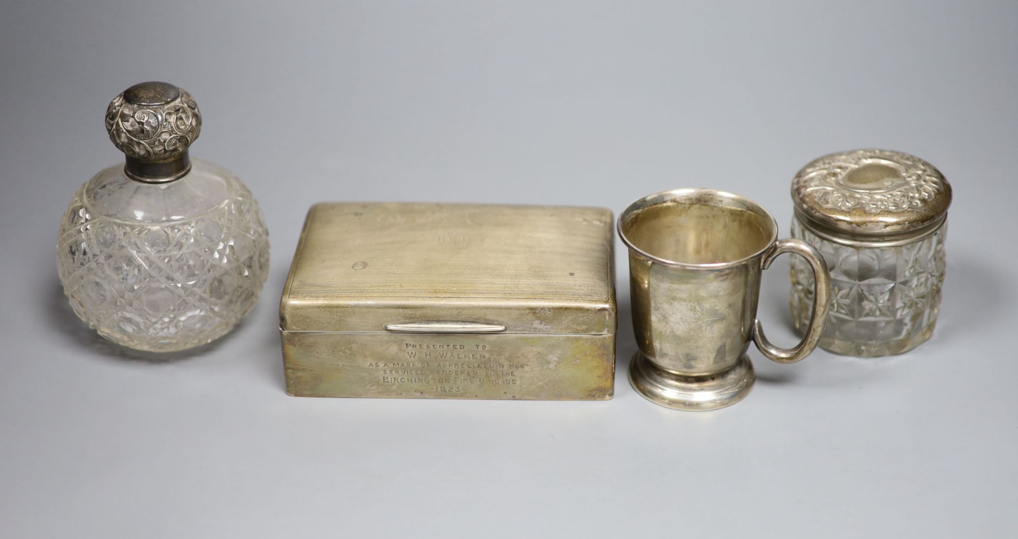 A George VI silver christening mug, a silver mounted cigarette box, a silver mounted scent bottle and toilet jar.